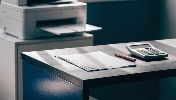 Ofis masasında hesap makinesi ve belgeler, arka planda çok fonksiyonlu yazıcı
